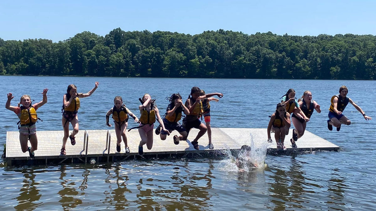 A group of children, wearing swimsuits and life jackets, are seen jumping off a dock and into the water.