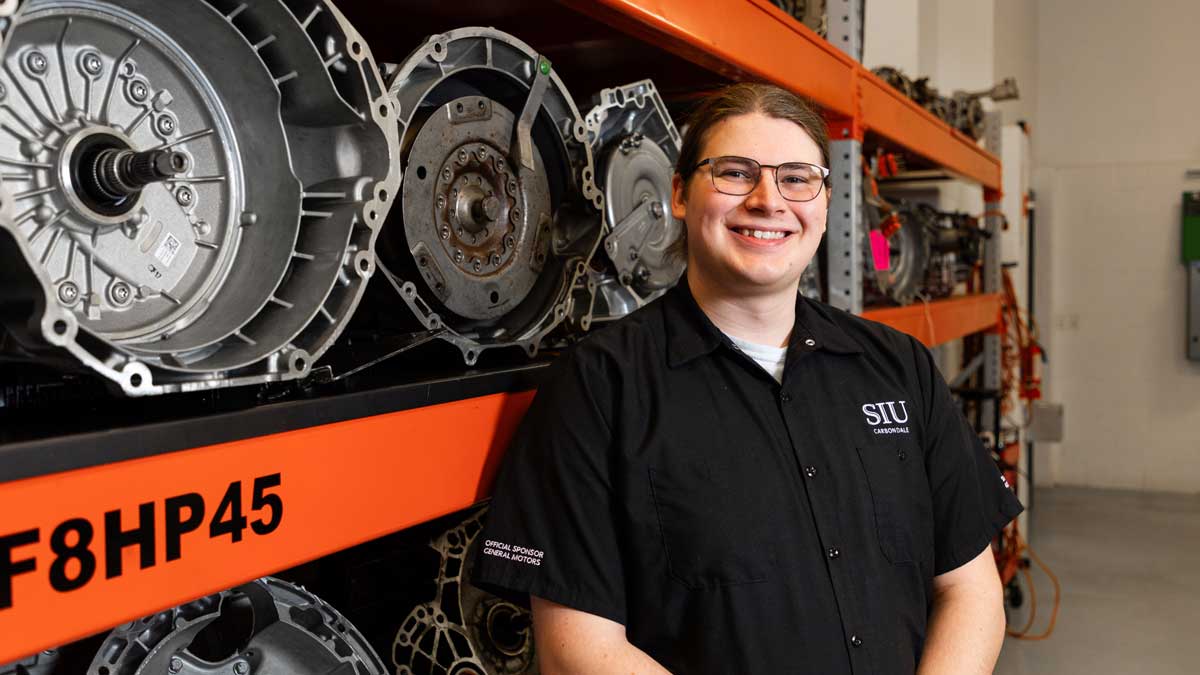 A young man, dark hair and glasses. Wearing a black button up shirt that says SIU Carbondale on the left chest. He is standing if front of racks that hold automotive transmissions